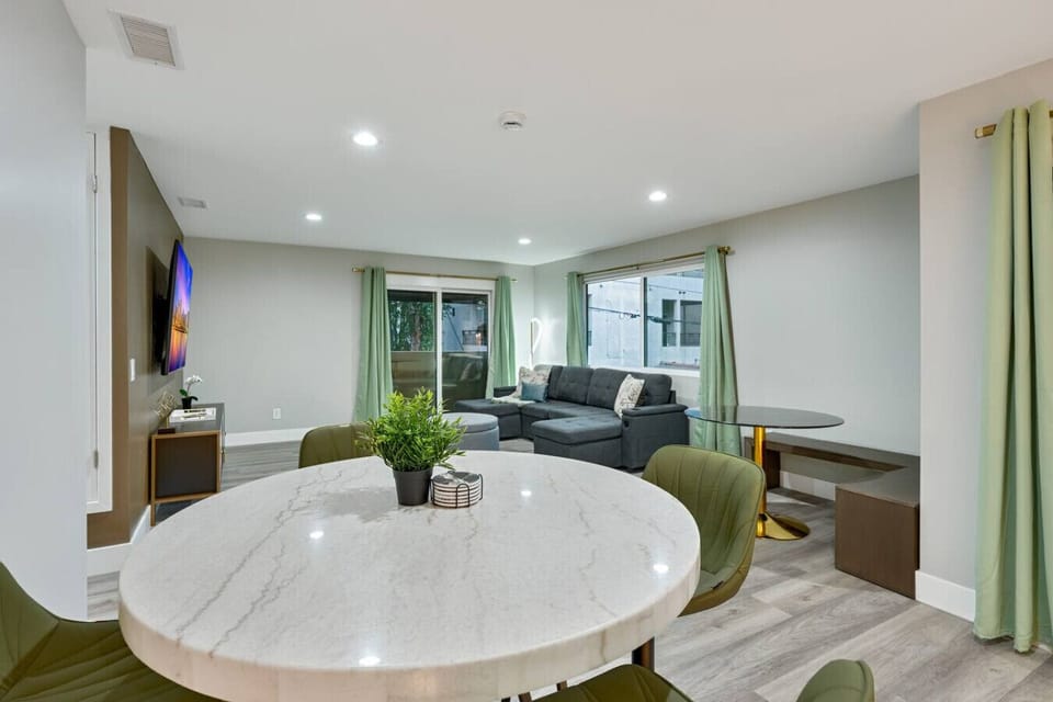 A marble dining table anchors this open living space, with the sofa and TV visible just beyond. Large windows bring in plenty of natural light throughout the day.