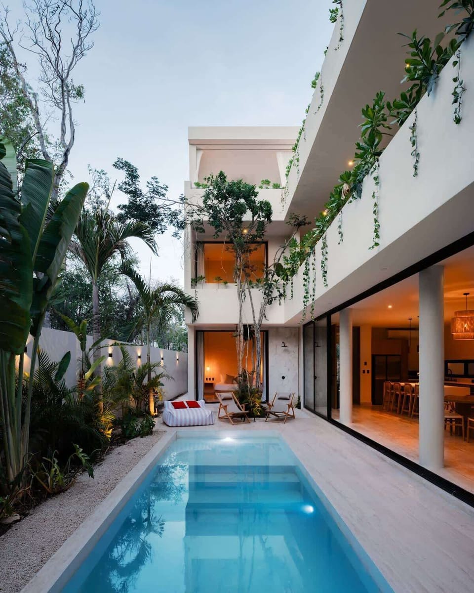 Two-story villa exterior at dusk with warm golden interior lighting and tropical trees, Tulum
