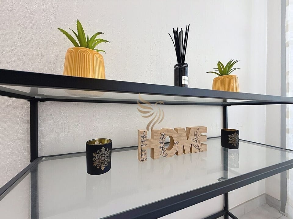 Installez-vous : une étagère noire aux accents bois et pots jaunes diffuse chaleur et parfum. Le mot `HOME` posé au centre invite à se sentir chez soi, entre bougies douces et lumière naturelle.