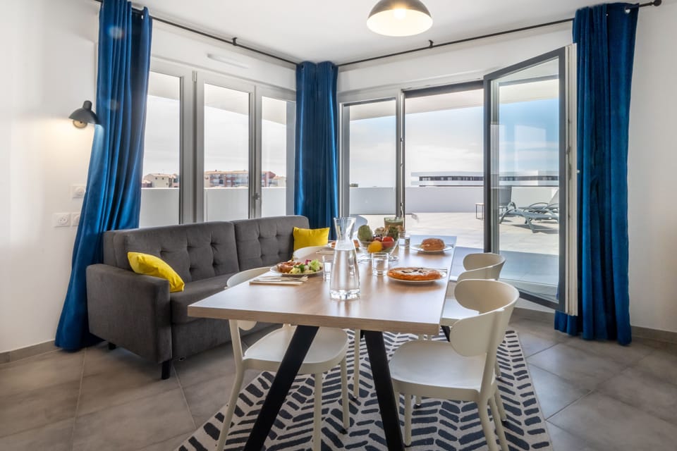 Dining table set for a meal, with a gray sofa and yellow accent cushion — natural light fills the room through blue-curtained glass doors.