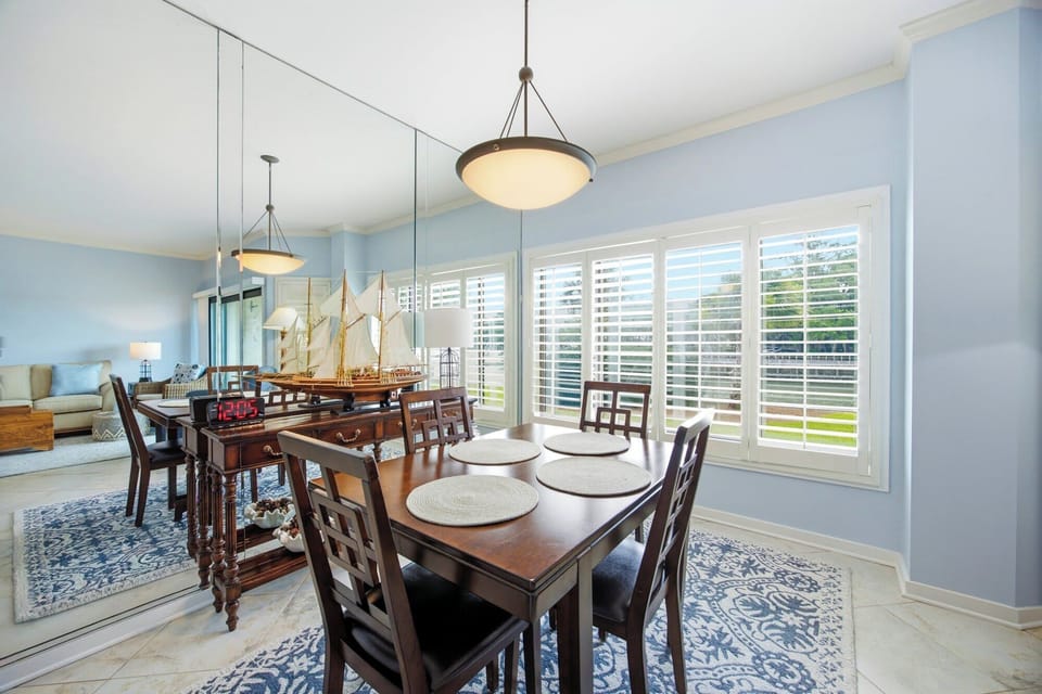 Gleaming dark wood furniture anchors this airy dining nook, where mirrored walls expand the coastal glow from white plantation shutters. Pale blue walls and a detailed model ship evoke a peaceful maritime charm for your morning coffee.