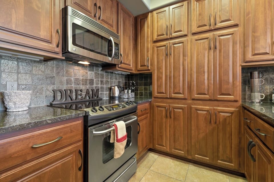 A close-up view of a kitchen workspace emphasizing an integrated stainless steel microwave above a modern stove, surrounded by abundant wooden cabinetry and a grey stone tile backsplash.