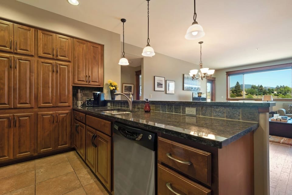 A view over the kitchen counter featuring a polished dark granite breakfast bar and a deep sink setup, looking out toward the dining and living spaces with a large window in the background.