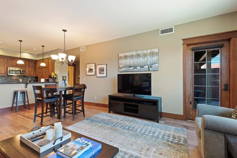 An open-concept living area with hardwood floors, showcasing a large wall-mounted flat-screen television on a dark media console, a patterned area rug, a glass door leading outside, and a kitchen bar in the background.