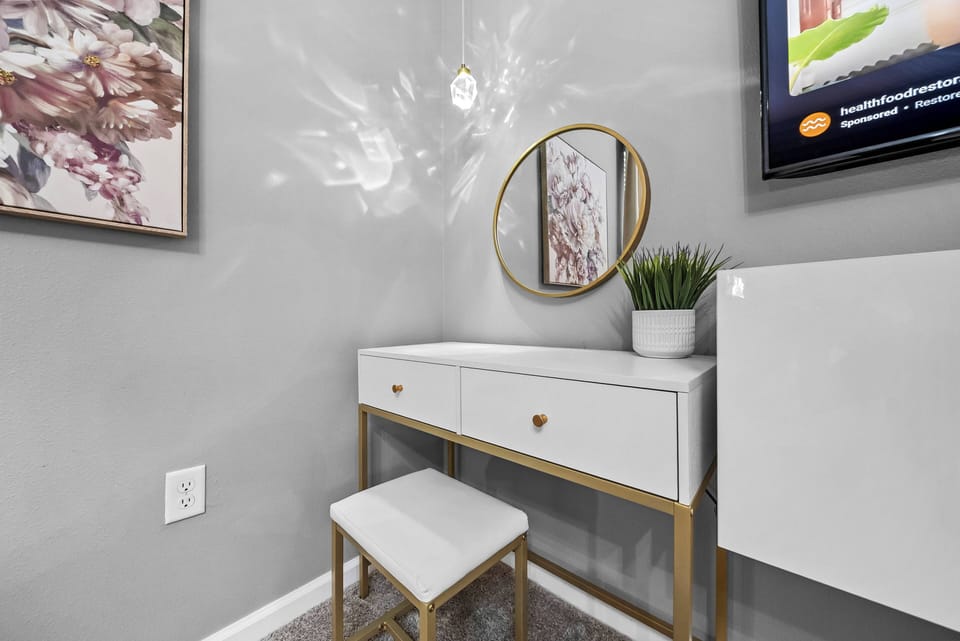 This chic vanity area boasts a sleek white desk with gold accents and a stylish round mirror. The soft gray walls and greenery create a calming atmosphere, making it the perfect spot to get ready for your day.