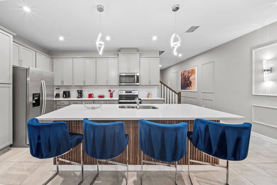 This stylish kitchen boasts a spacious island with sleek white countertops and vibrant blue bar stools for casual dining. Modern appliances and soft gray cabinetry create a bright, inviting atmosphere for your culinary adventures.
