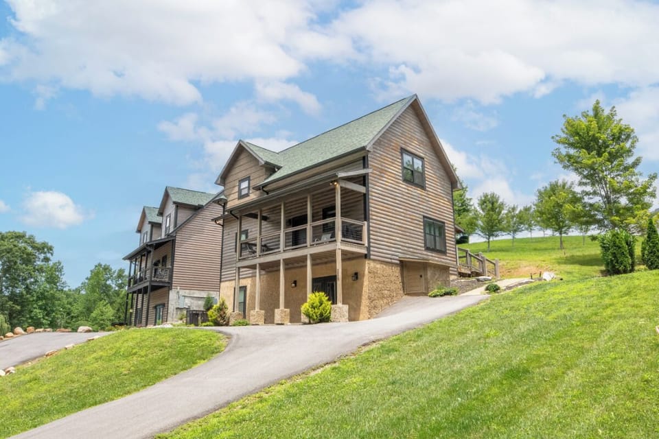 A spacious hillside property featuring natural wood siding and multiple levels of covered deck space, surrounded by lush lawns and mature trees under a bright blue sky.