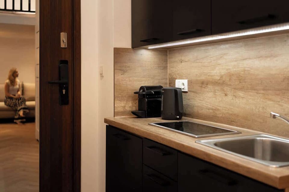 W apartamencie znajduje się również funkcjonalny aneks kuchenny, wyposażony w płytę kuchenną, czajnik elektryczny oraz ekspres do kawy, umożliwiający przygotowanie prostych posiłków i porannej kawy. Obok aneksu usytuowano stół jadalniany z krzesłami.