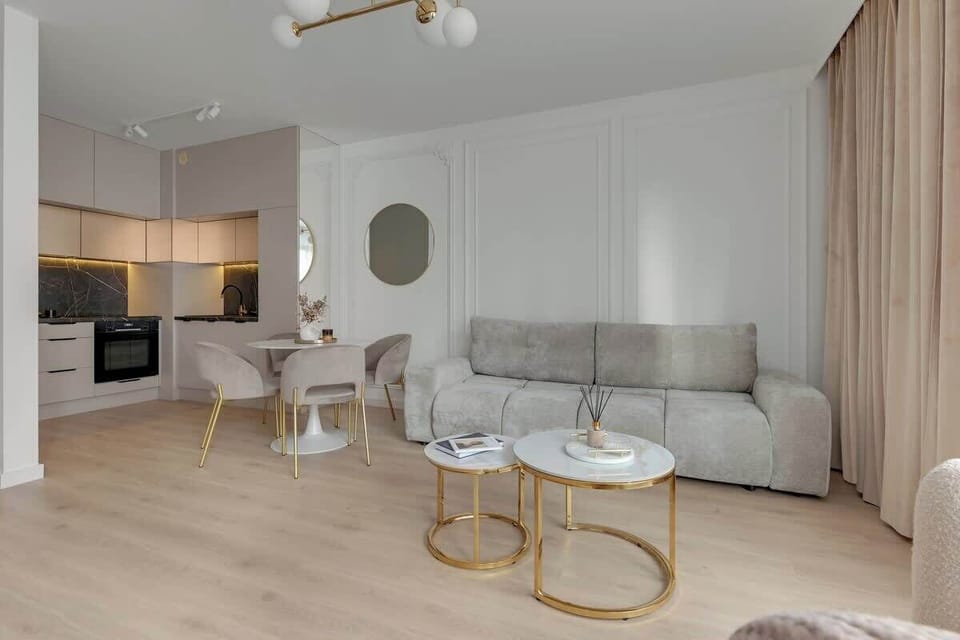 Przestronny salon z rozkładaną sofą i dużymi oknami zapewnia komfortowy wypoczynek w jasnym, pełnym światła wnętrzu. Elegancka jadalnia z marmurowym stołem i welurowymi krzesłami dopełnia całość.