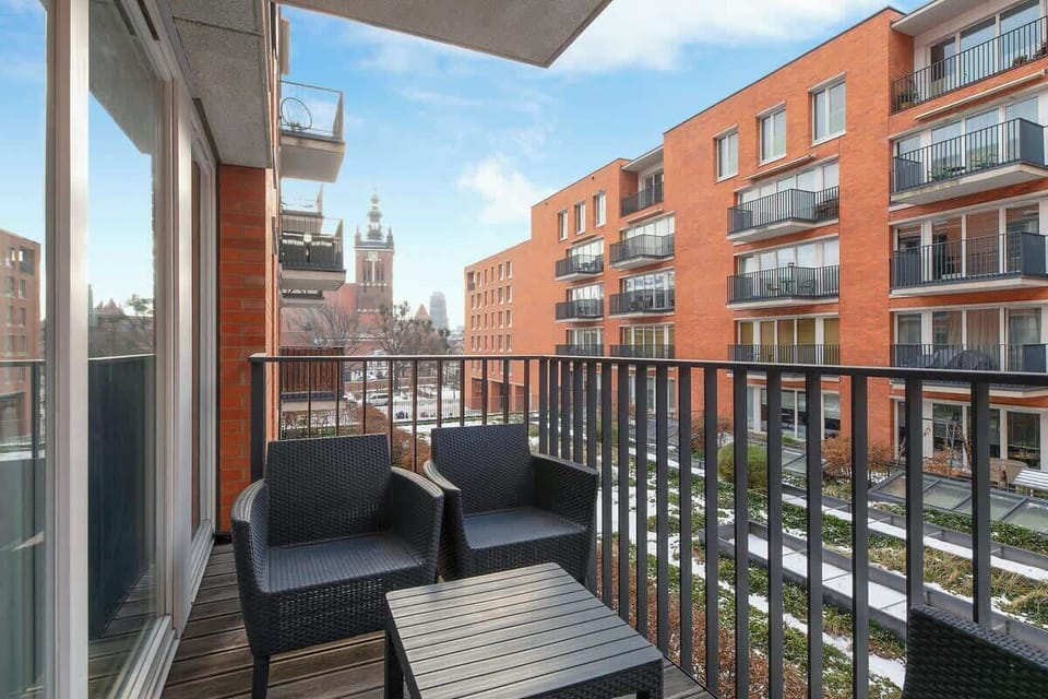Z pokoju dziennego prowadzi wyjście na balkon z komfortowymi fotelami, skąd rozciąga się widok na zielone patio i ogródki, tworząc idealne miejsce na poranną kawę lub wieczorny odpoczynek.