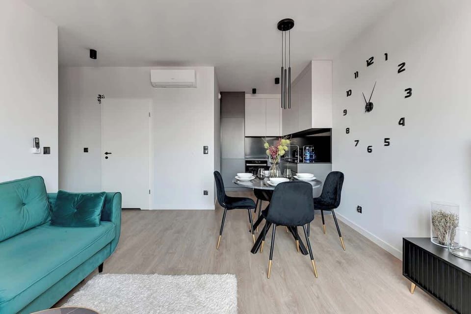 Nowoczesny salon z aneksem kuchennym urządzony w jasnych barwach. Turkusowa sofa, stolik kawowy i miękki dywan tworzą strefę relaksu. Obok znajduje się jadalnia oraz minimalistyczna kuchnia, a duże okno dobrze doświetla wnętrze.