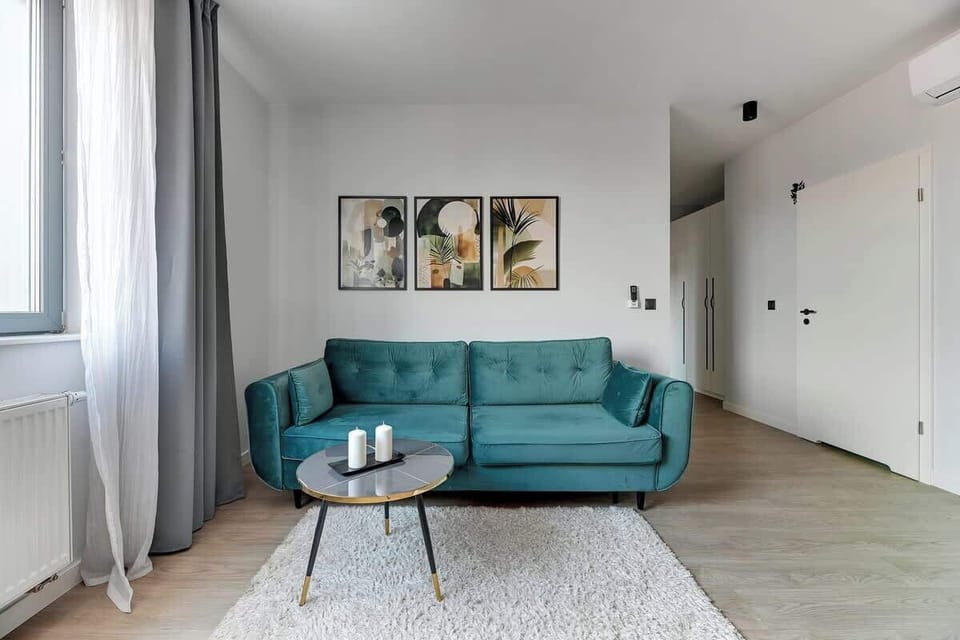 Nowoczesny salon z aneksem kuchennym urządzony w jasnych barwach. Turkusowa sofa, stolik kawowy i miękki dywan tworzą strefę relaksu. Obok znajduje się jadalnia oraz minimalistyczna kuchnia, a duże okno dobrze doświetla wnętrze.