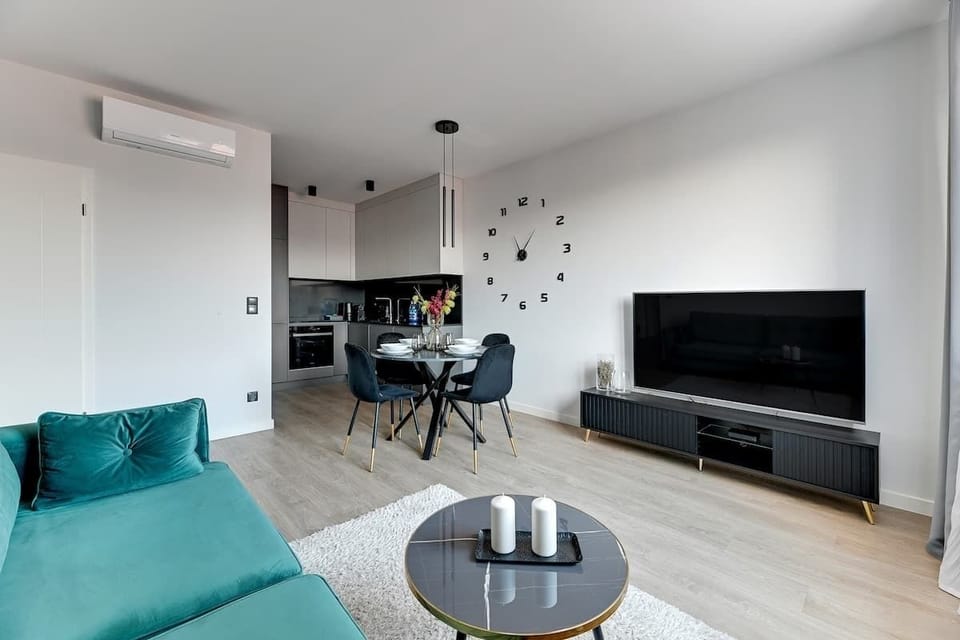 Nowoczesny salon z aneksem kuchennym urządzony w jasnych barwach. Turkusowa sofa, stolik kawowy i miękki dywan tworzą strefę relaksu. Obok znajduje się jadalnia oraz minimalistyczna kuchnia, a duże okno dobrze doświetla wnętrze.