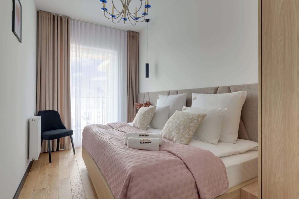 Przytulna sypialnia urządzona w eleganckim, nowoczesnym stylu zachwyca jasnym wnętrzem, naturalnym drewnem i subtelnymi dodatkami. Duże łóżko oraz dostęp do balkonu tworzą komfortową przestrzeń idealną do wypoczynku i relaksu.