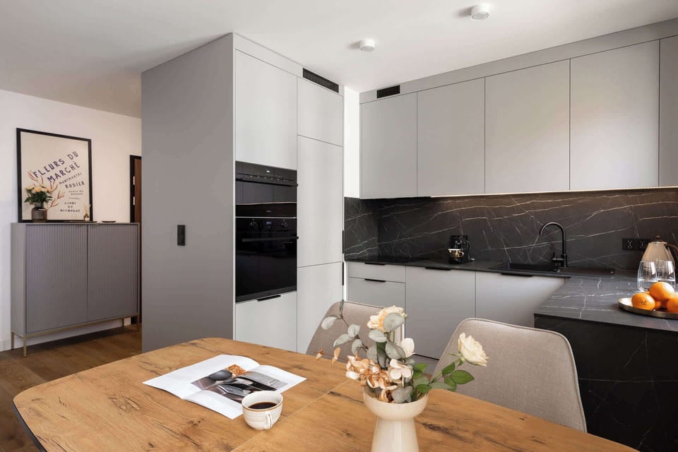 Apartament posiada w pełni wyposażony aneks kuchenny, utrzymany w minimalistycznym, nowoczesnym stylu. Jasne fronty szafek, ciemny blat oraz elegancki kamienny motyw nad blatem tworzą spójną i estetyczną całość.