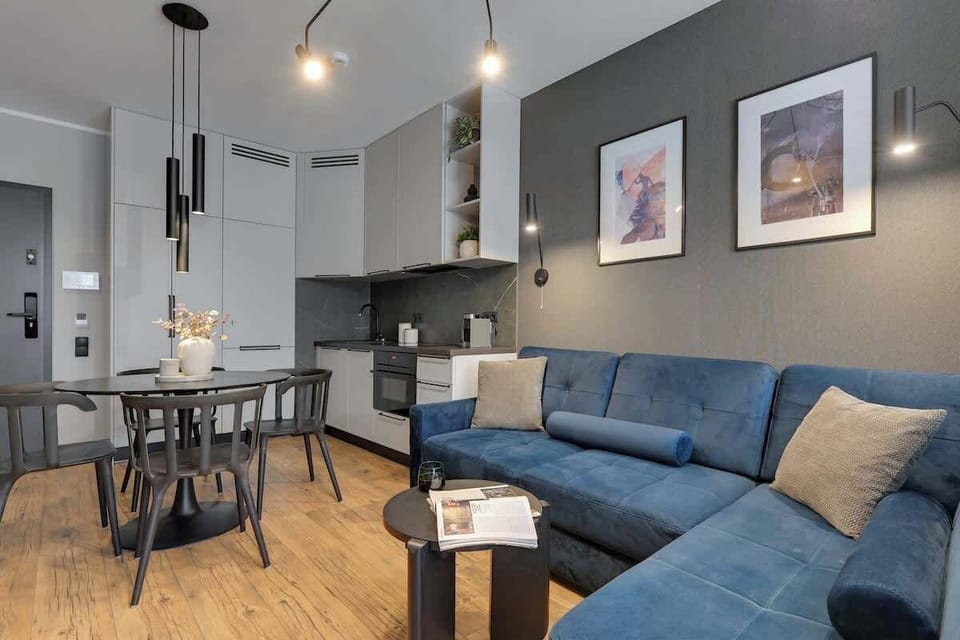 Stylowy salon z aneksem kuchennym i częścią jadalnianą, urządzony w nowoczesnym, eleganckim stylu. Komfortowa sofa, designerskie oświetlenie oraz w pełni wyposażona kuchnia tworzą idealną przestrzeń do relaksu i codziennego użytkowania.