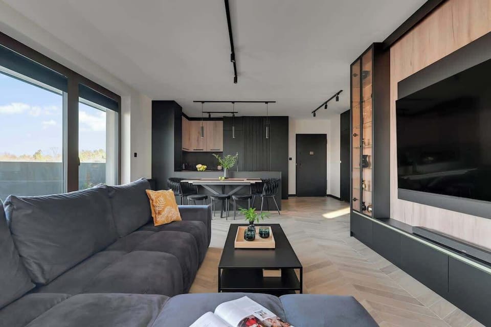 Salon utrzymany jest w nowoczesnej, minimalistycznej stylistyce, w której dominują ciemne kolory, naturalne drewno oraz oświetlenie LED. Znajduje się tu duża, rozkładana sofa, a naprzeciw niej zamontowano telewizor z kanałami TV oraz Smart TV.