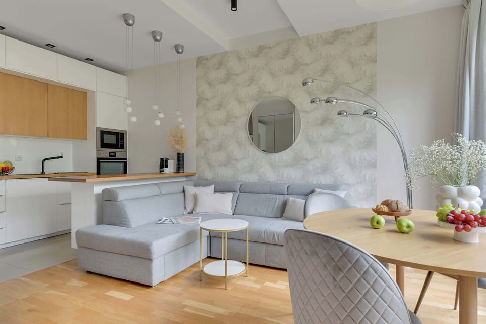 Nowoczesny salon z dużymi przeszkleniami i widokiem na zieleń tworzy jasną oraz komfortową przestrzeń do wypoczynku. Stylowe wnętrze łączy przytulną część wypoczynkową z funkcjonalnym aneksem kuchennym i jadalnią, zapewniając idealne warunki zarówno 