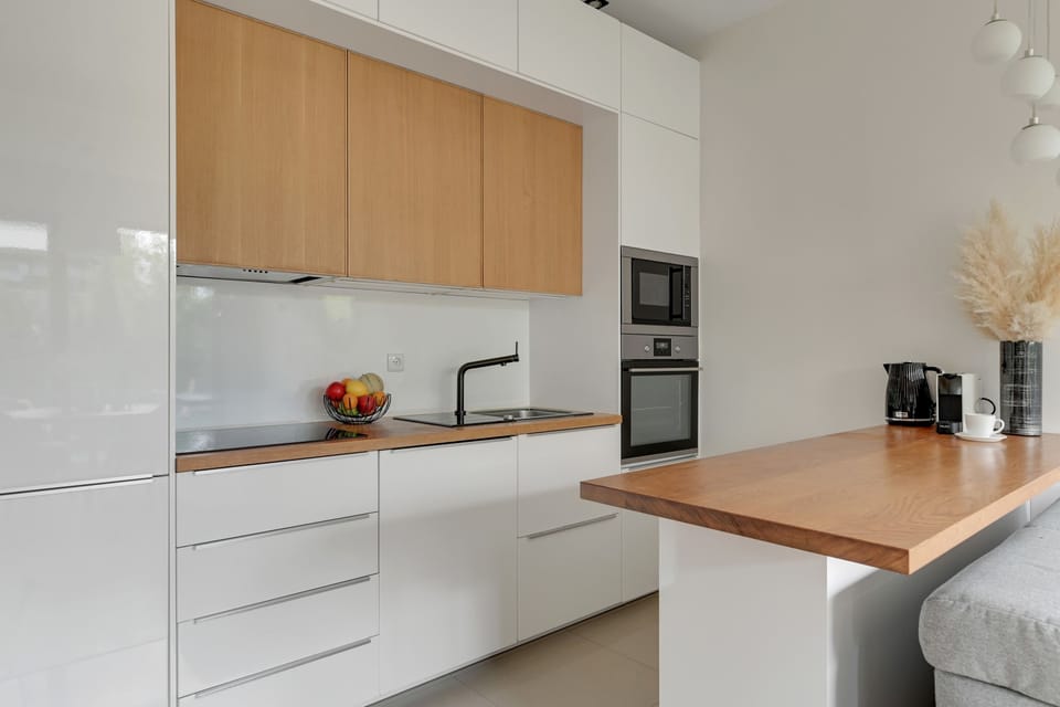 Nowoczesna kuchnia została wyposażona w wysokiej klasy sprzęty AGD oraz funkcjonalną zabudowę w jasnych kolorach z drewnianymi akcentami. Minimalistyczny design i przestronny blat tworzą komfortową przestrzeń do gotowania oraz wspólnego spędzania cza