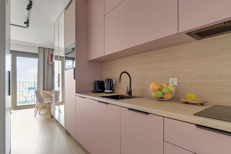 Stylowy aneks kuchenny w pudrowych odcieniach łączy nowoczesny design z funkcjonalnością. Jasne drewniane akcenty, pełne wyposażenie oraz minimalistyczne detale tworzą przytulną i elegancką przestrzeń.