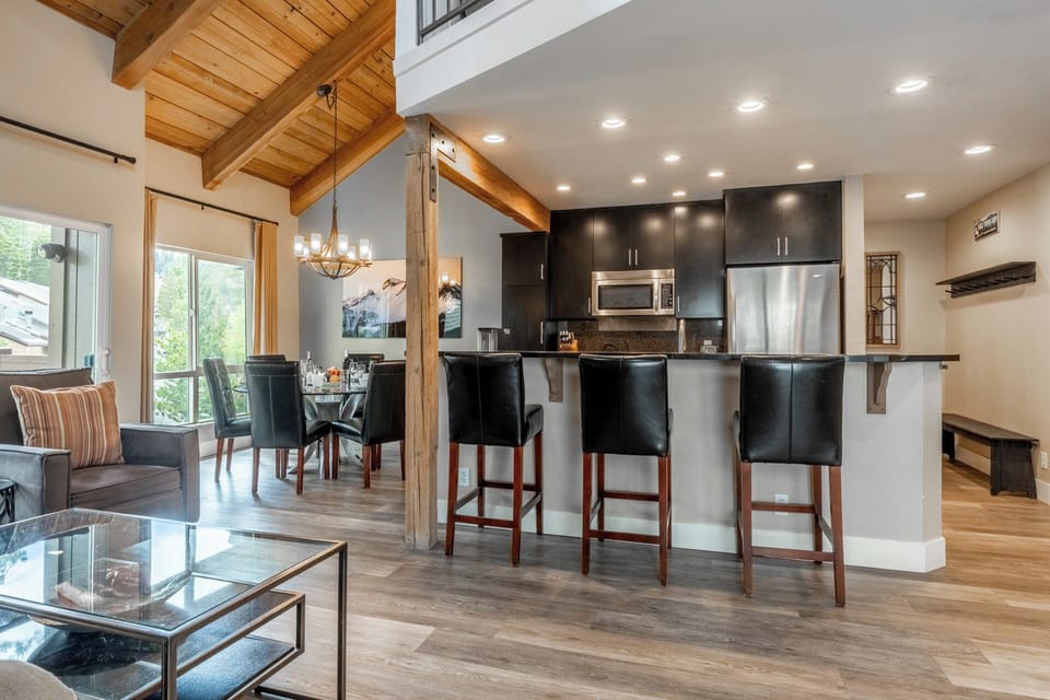 Your open-concept living space features soaring wood-beam ceilings, a sleek modern kitchen with breakfast bar, and abundant natural light streaming through large windows.