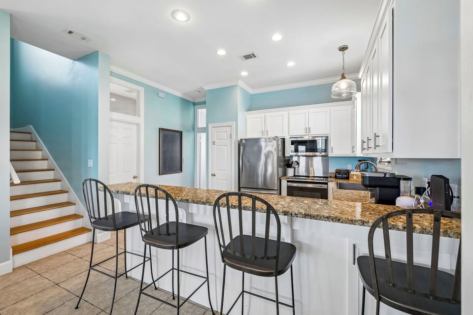 Your fully equipped kitchen features modern stainless steel appliances, a spacious granite island with seating for four, and bright, coastal-inspired blue walls that create a fresh, welcoming atmosphere.