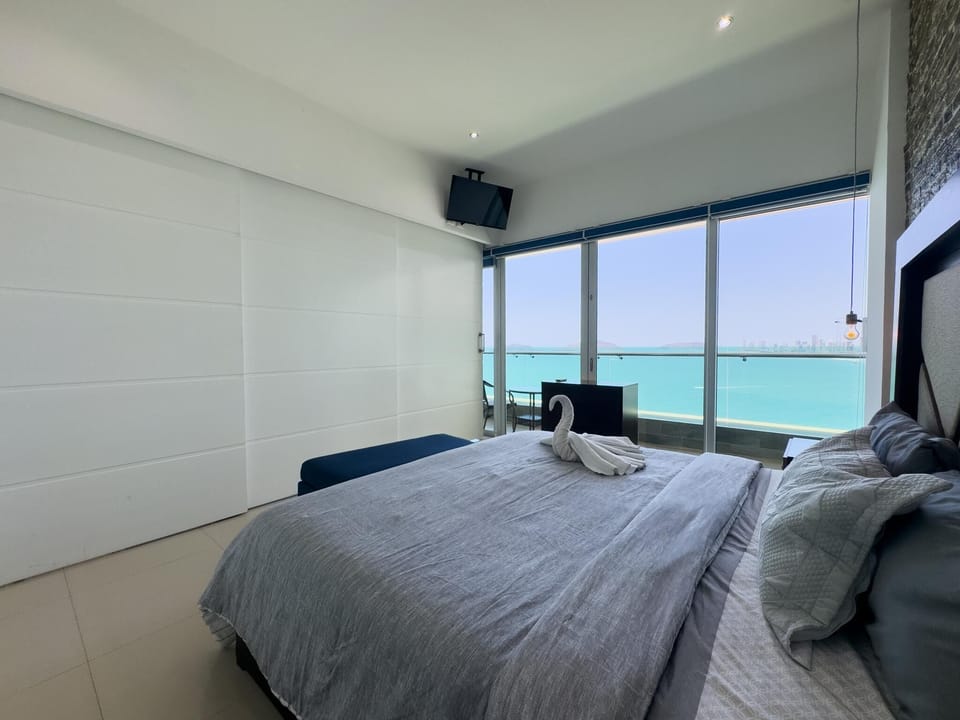 Bedroom features a gray quilted bed with towel swan, navy bench, and sliding glass doors overlooking turquoise ocean.