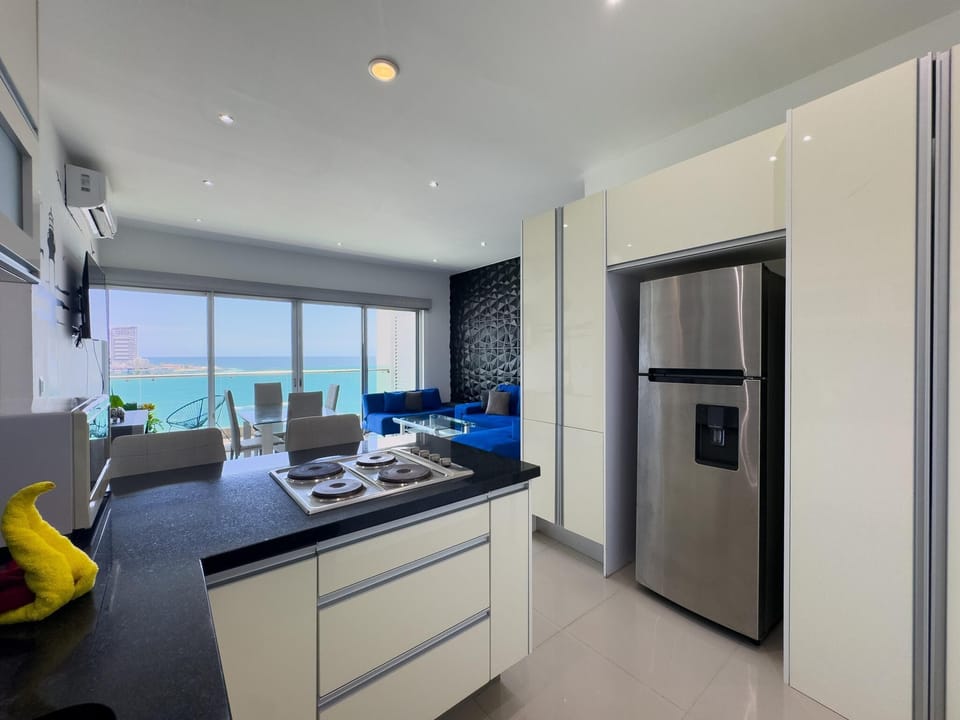 Glossy cream cabinetry surrounds a stainless steel refrigerator beside a black granite island with electric cooktop.