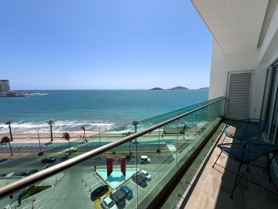 Balcony with two teal woven chairs overlooks the ocean, beach, and street below through glass railing.