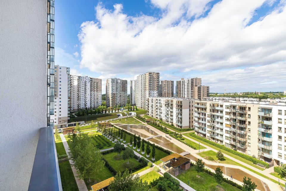 A high-angle view from a balcony looks out over a beautifully landscaped modern residential courtyard. Green lawns, walkways, and contemporary apartment blocks extend under a bright blue sky with white clouds.