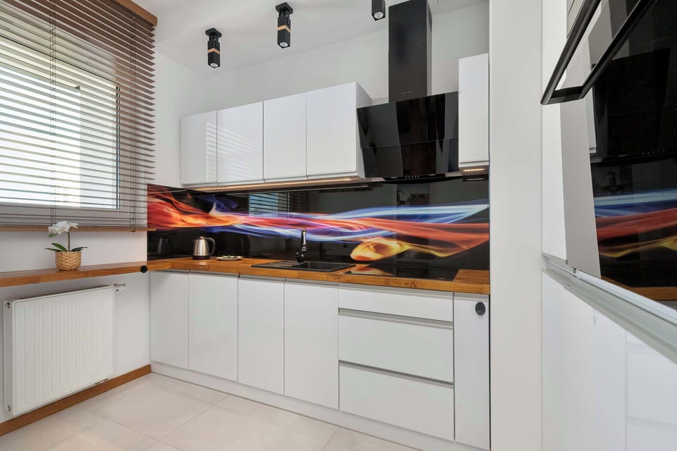 A modern kitchen boasts glossy white cabinetry paired with a warm wooden countertop and an eye-catching colorful abstract backsplash design. A sleek black cooker hood stands out against the white walls.