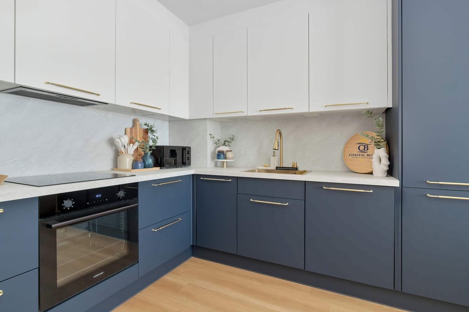 A modern kitchen showcases white upper cabinets and navy blue lower cabinets. It includes high-end built-in appliances like an oven and a induction stovetop, set against a light wood floor.