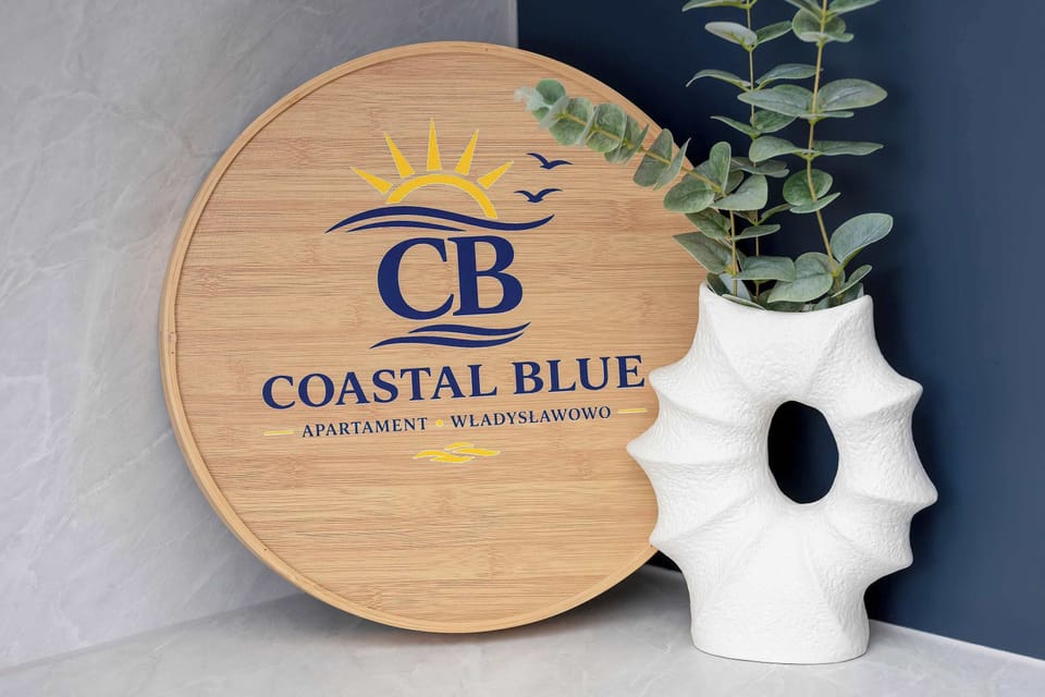 A decorative arrangement features a round wooden sign with a sun and wave logo next to a white textured vase. Green eucalyptus stems emerge from the vase against a deep blue backdrop.