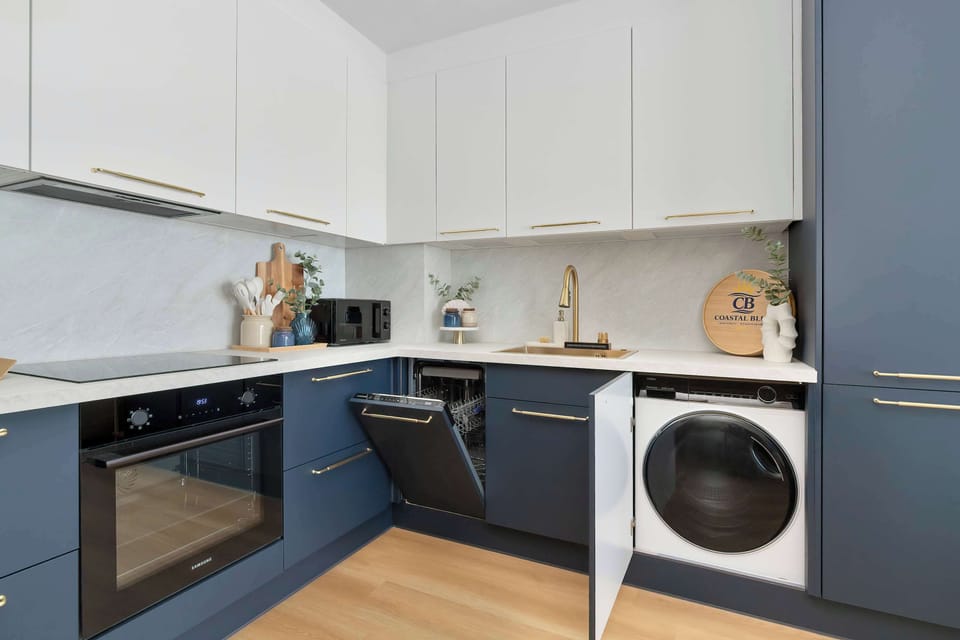 The kitchen is shown with several lower cabinet doors open, revealing a built-in dishwasher and a washing machine. This showcases the functional, space-saving appliances included in the layout.