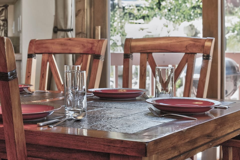 Picture yourself enjoying meals at this inviting wooden table, where warm natural light streams in through large windows overlooking a lush green garden setting.