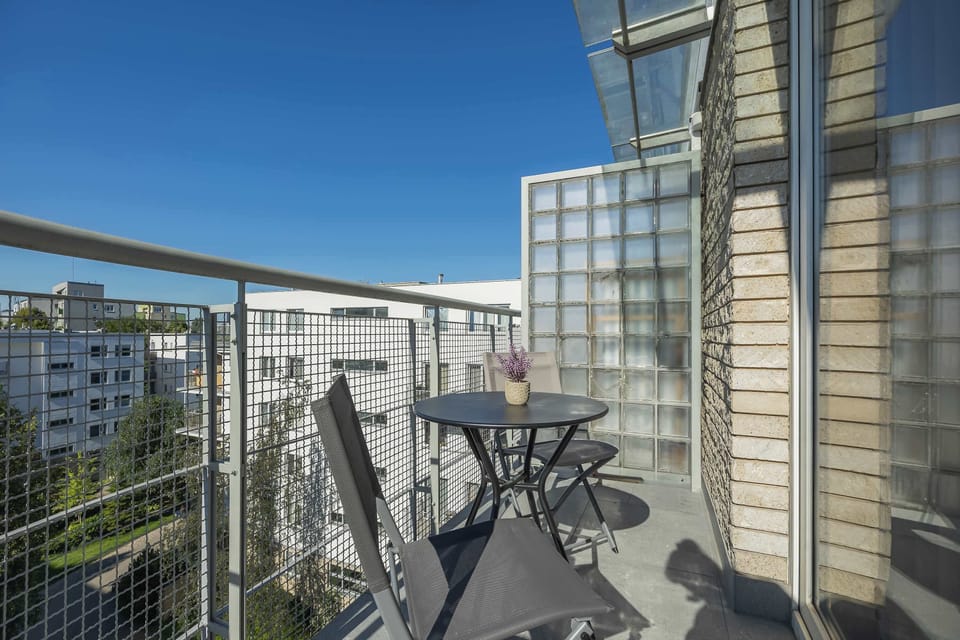 A private balcony offers a small bistro table and chairs overlooking nearby apartment buildings. The elevated position provides an open urban view.