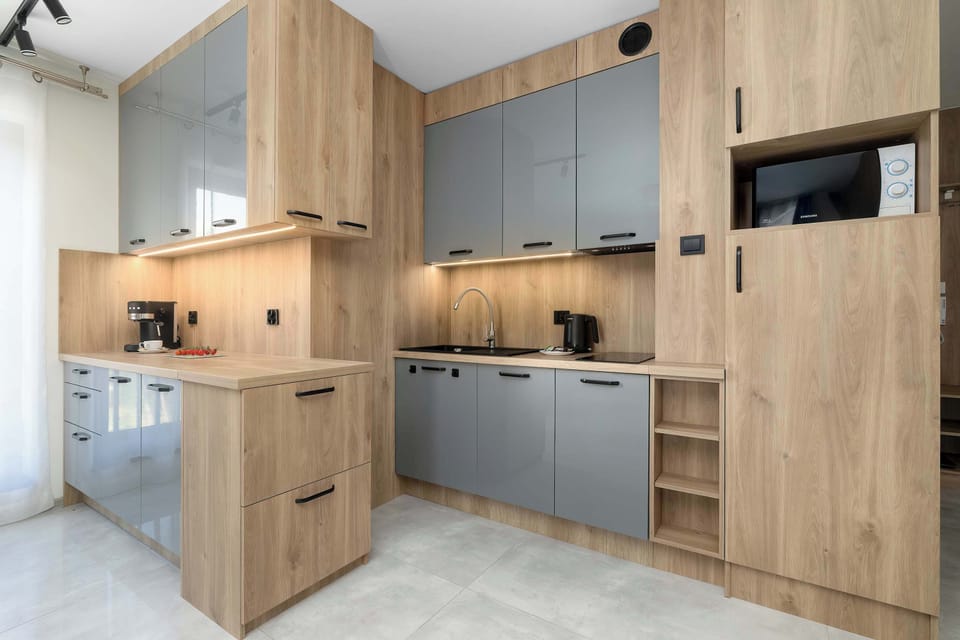 A modern kitchen combines grey minimalist cabinet fronts with warm wooden elements. Built-in appliances are neatly fitted into the layout.