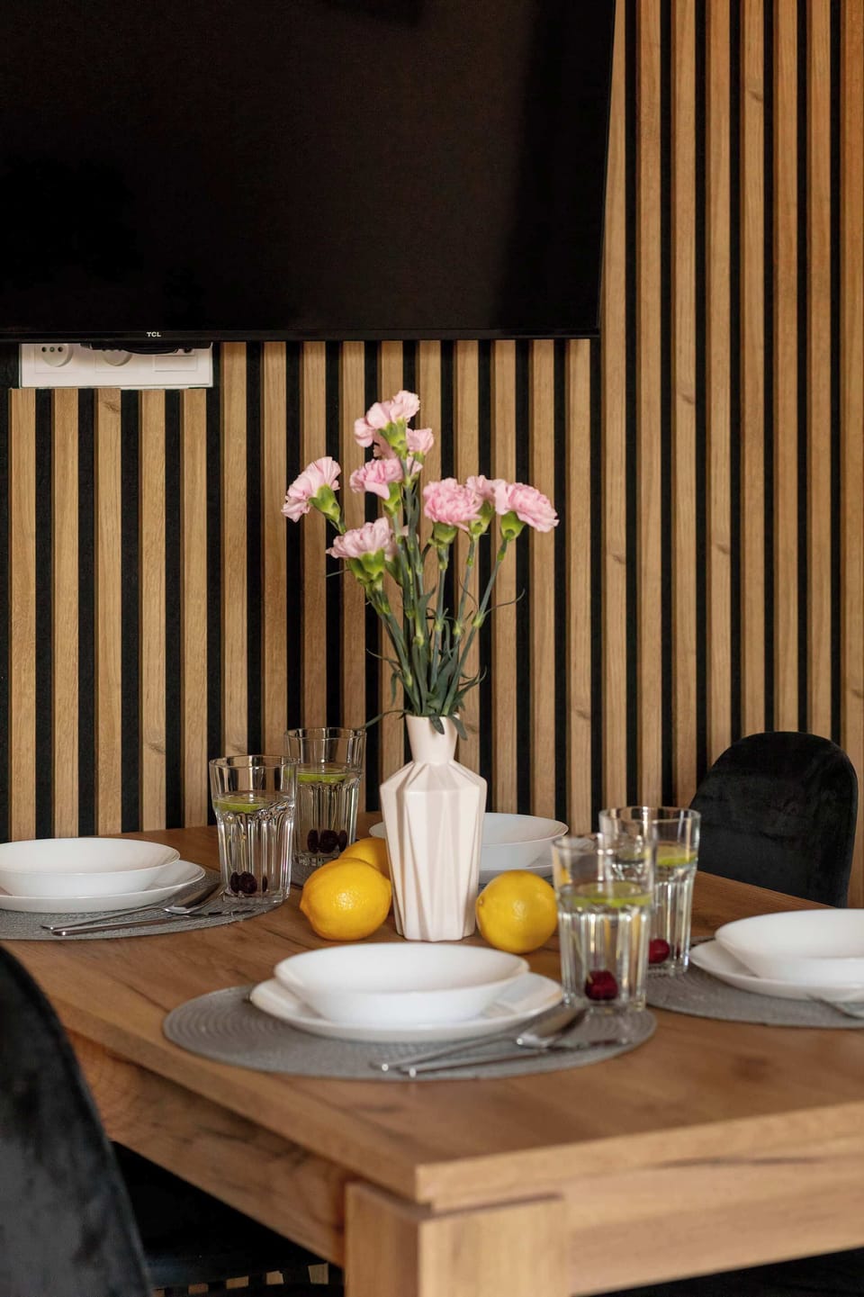 A close-up of the wooden dining table reveals a vase with pink flowers and fresh lemons. The background features a stylish slatted wood wall.