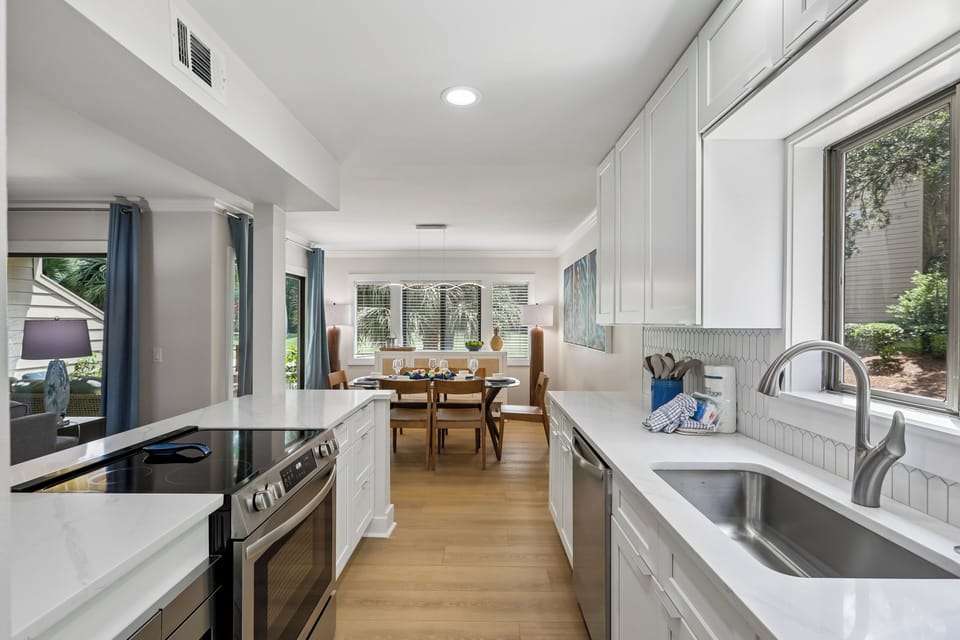 Bright, modern kitchen with sleek white cabinetry, marble countertops, and stainless steel appliances. Large windows overlook the garden, filling the space with natural light for inspiring meal prep.