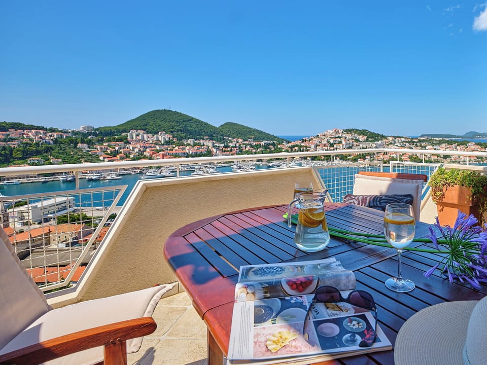 Relax on the sunlit balcony with panoramic harbor views. A wooden table and refreshing drinks await your peaceful retreat.