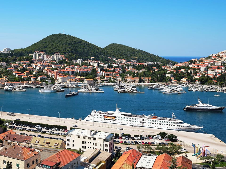 Enjoy panoramic views over Gruž Harbor. Sparkling blue waters, a docked cruise ship, and lush hills create a scenic backdrop.