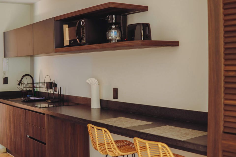 Fully-equipped modern kitchenette. Dark wood cabinetry and countertops feature an induction cooktop and sink. A floating shelf conveniently holds a microwave, drip coffee maker, toaster, and bottle of water. Woven stools complete the breakfast bar.