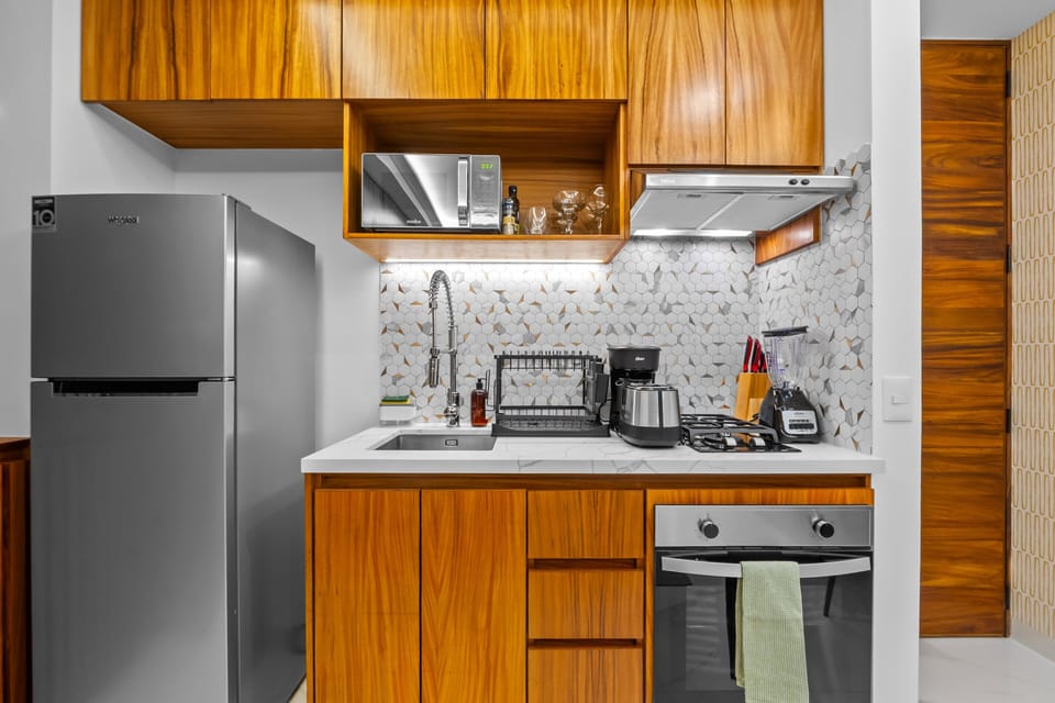 Features a stainless steel refrigerator, built-in oven, gas cooktop, microwave, and blender. Styled with rich wood cabinetry, a chic geometric tile backsplash, and a deep sink with a pull-down faucet.