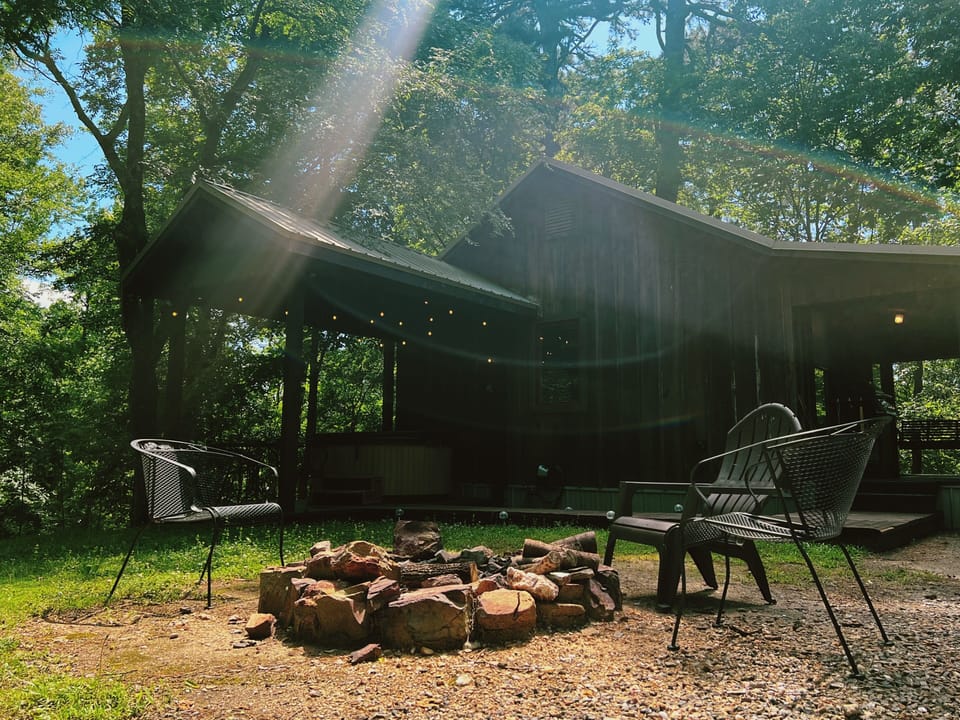 Imagine evenings gathered around this stone fire pit, nestled beneath towering trees. The cabin's covered porch glows warmly with string lights, inviting you to unwind in cozy outdoor seating.