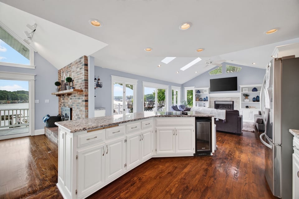 Sunlit lakeside living with an open kitchen and living area featuring gleaming hardwood floors, vaulted ceilings, and wall-to-wall windows framing breathtaking water views.