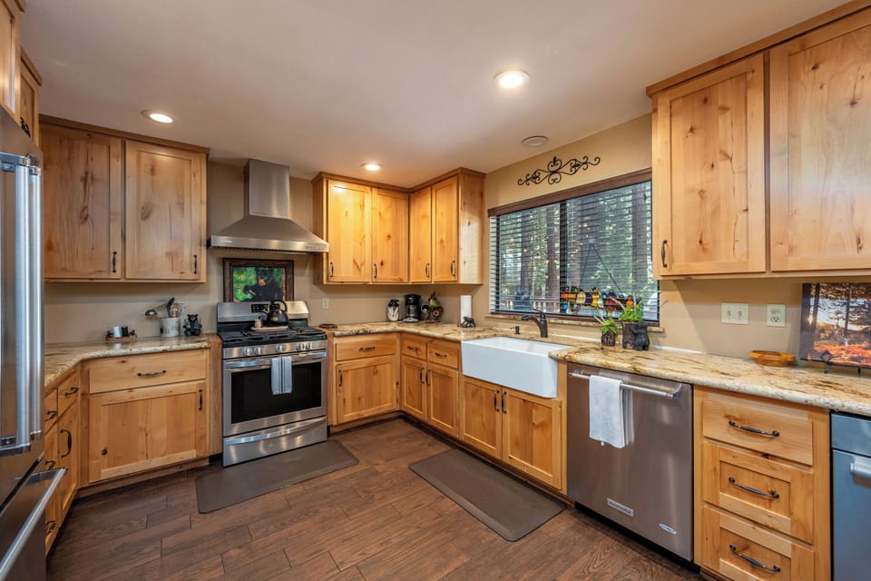 Your gourmet kitchen features warm knotty pine cabinetry, stainless steel appliances, and a spacious farmhouse sink—perfect for preparing meals with a view of the forest through the window.