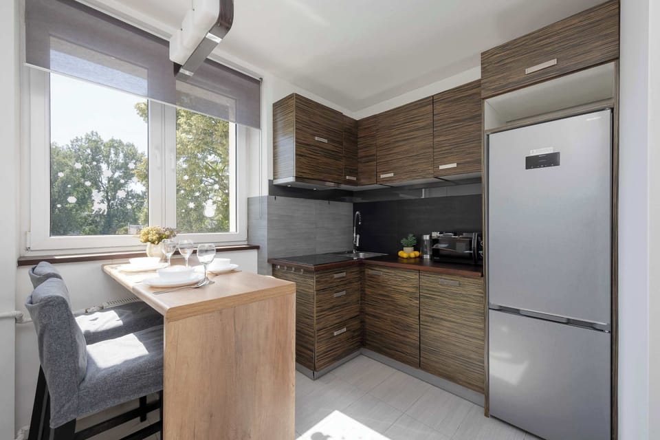A fully equipped kitchen with wooden cabinets, black countertops, and a dining table by the window. Large windows provide plenty of natural light.