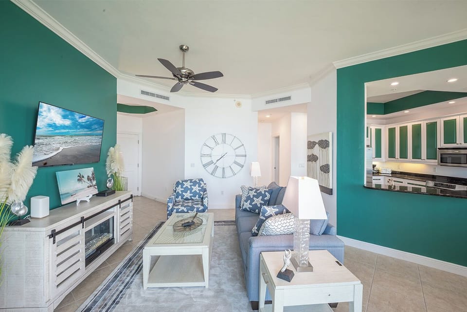 Bright, open-concept living room and kitchen area features teal accent walls, coastal-themed decor, and a mounted flat-screen TV.