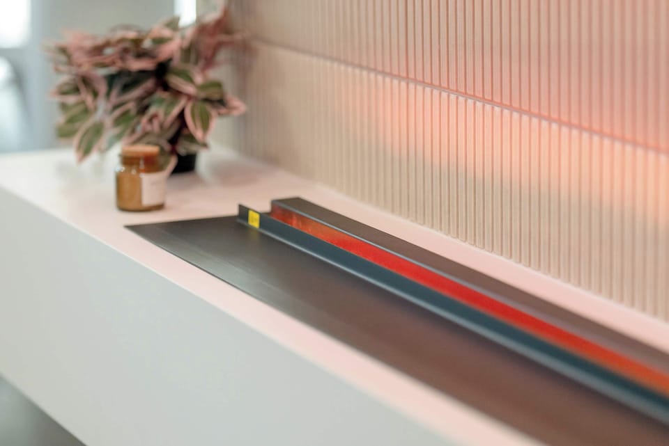 A close-up view captures a sleek white countertop with a long, narrow built-in bioethanol fireplace containing decorative logs. The soft orange glow introduces warmth to the minimalist interior.