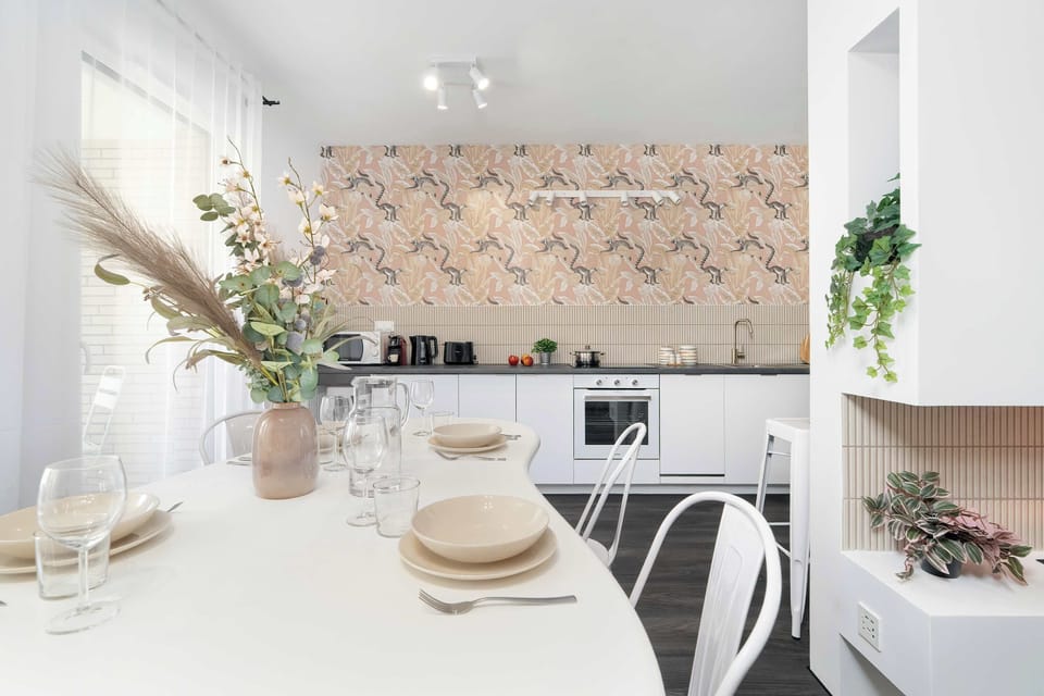 A white oval dining table is set with elegant dinnerware and a decorative vase filled with dried pampas grass. In the background, the modern kitchen and its distinctive wall pattern are visible.